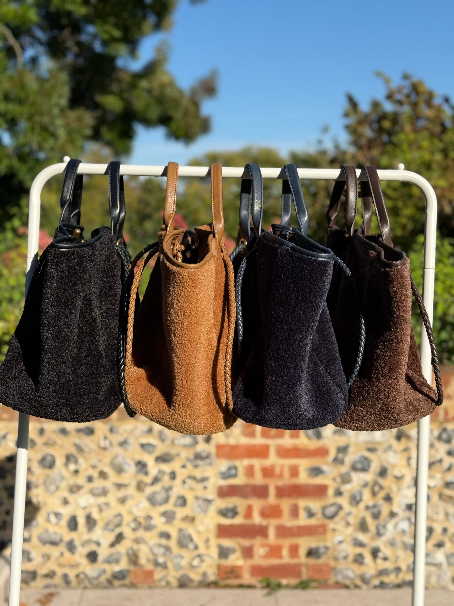 Five handbags of different colors hanging on a rack outdoors with a brick wall and greenery in the background.